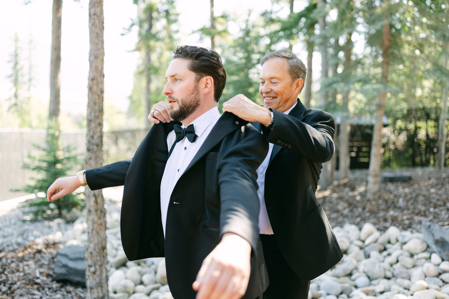 An older man helps a younger man in a tuxedo adjust his bow tie in a forest setting