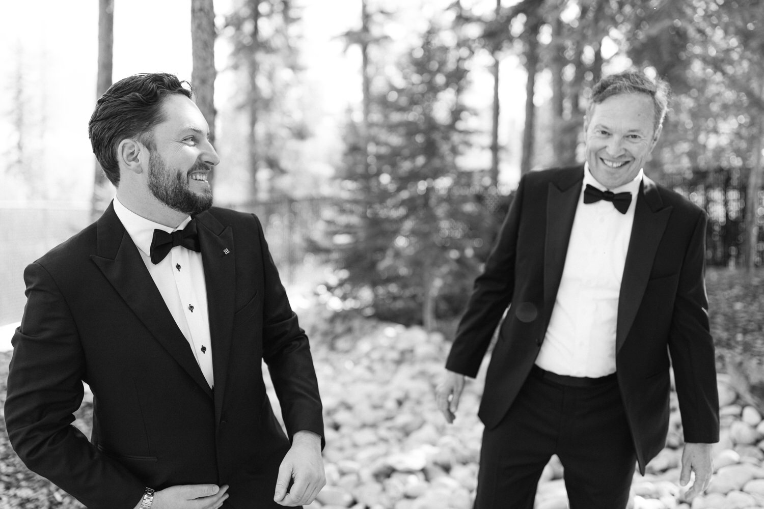 Two smiling men in tuxedos enjoying a moment in a forest setting, captured in a black and white photograph.