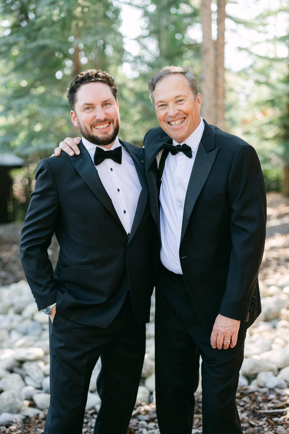 Two men smiling in formal black tuxedos with bow ties, standing in an outdoor setting with trees in the background.