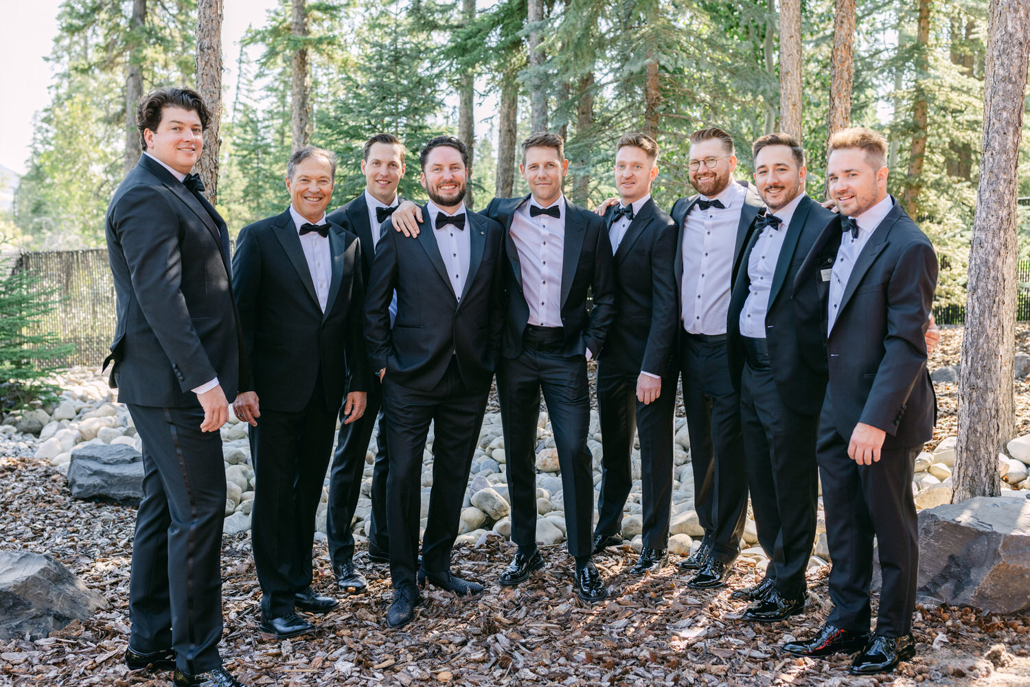 A group of men dressed in black tuxedos with bow ties and white shirts, smiling and standing together in an outdoor setting with trees and rocks in the background.