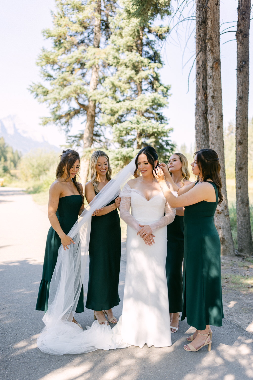 A bride in a white dress is being assisted by her four bridesmaids dressed in dark green, amid a natural setting with trees and mountains in the background.