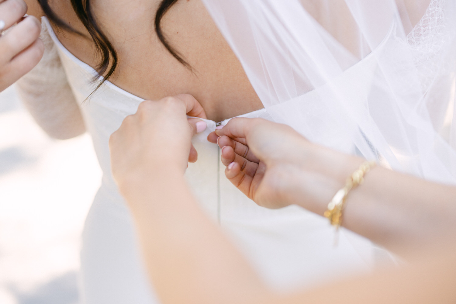 Close-up of a person fastening the back of a bride's dress, with focus on the hands and the white fabric