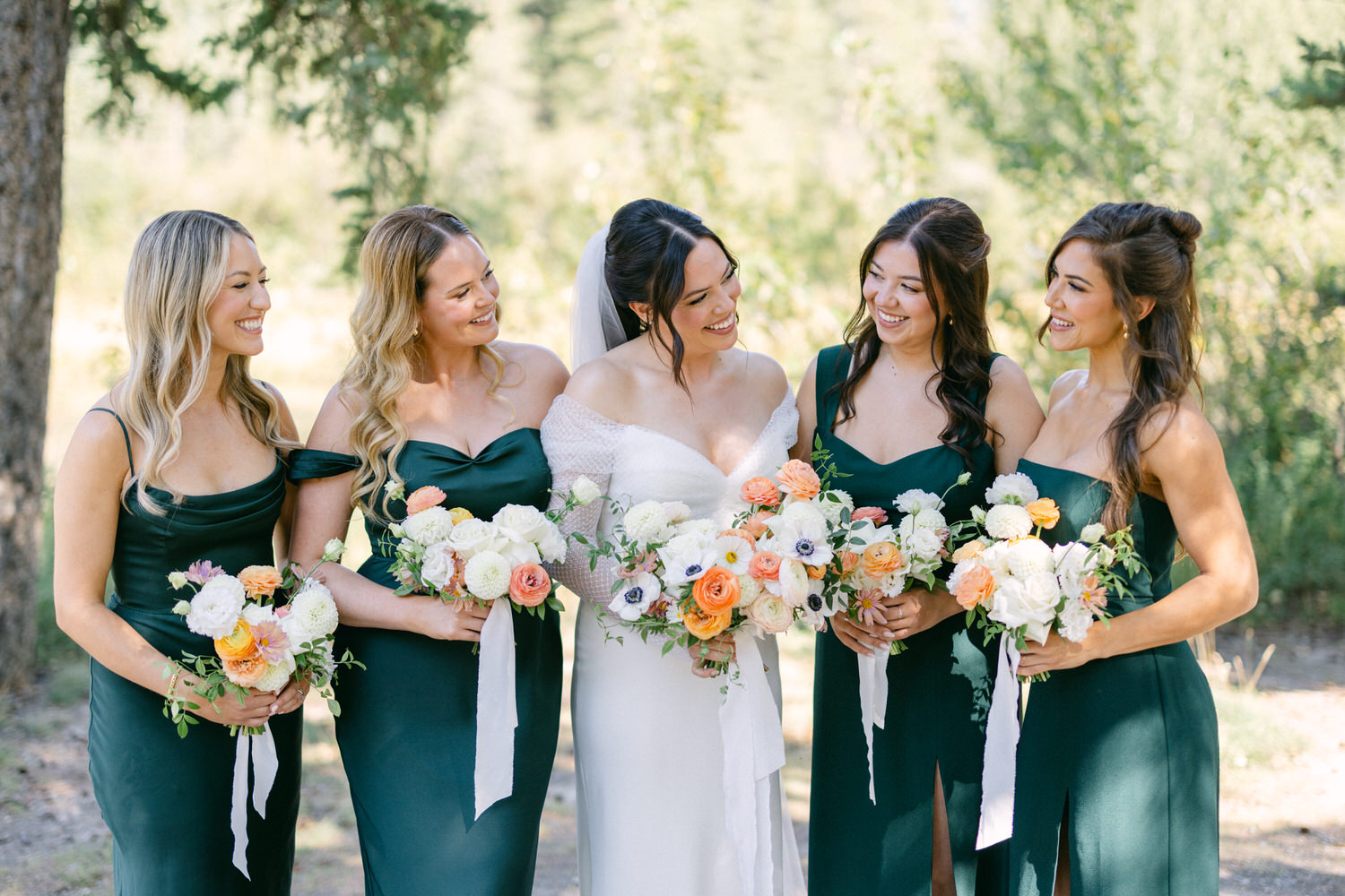 A bride in a white dress flanked by bridesmaids in dark green dresses, all holding bouquets and smiling outdoors.