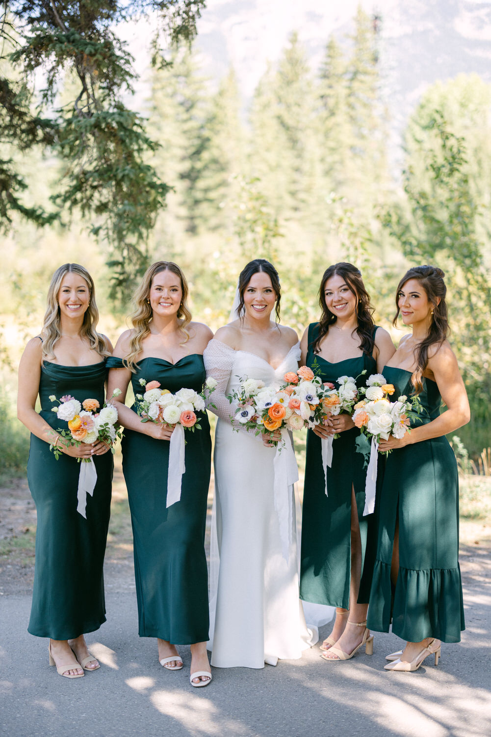 A bride in a white dress is flanked by her bridesmaids in dark green dresses, all holding bouquets, with a backdrop of trees.