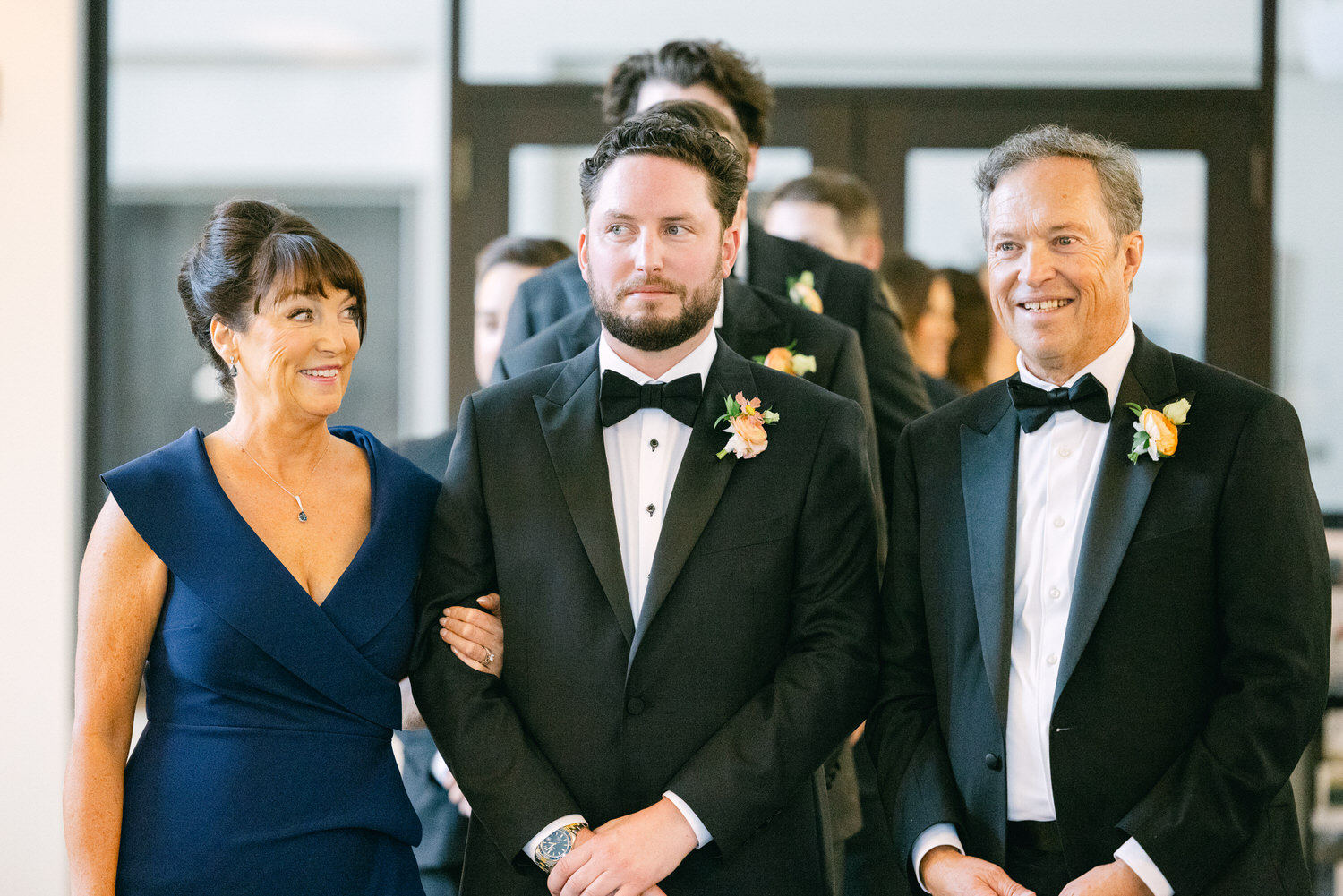 Three people, likely family members, dressed in formal attire at a wedding ceremony with joyous expressions.