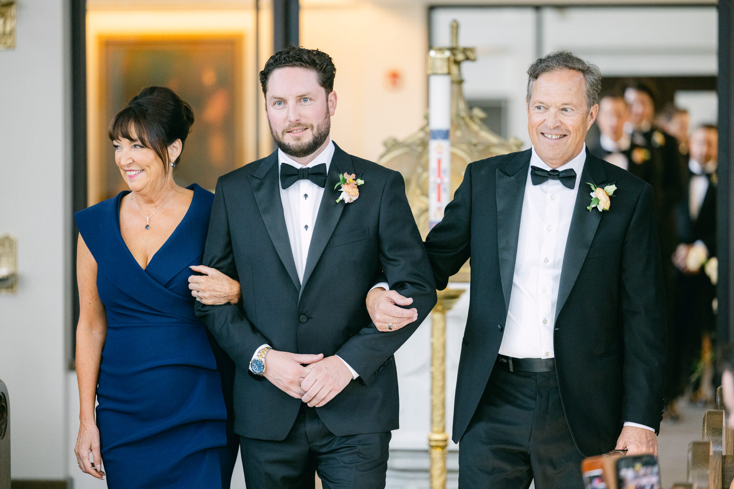 A woman and two men in formal attire walking down the aisle at a wedding ceremony, with onlookers in the background.