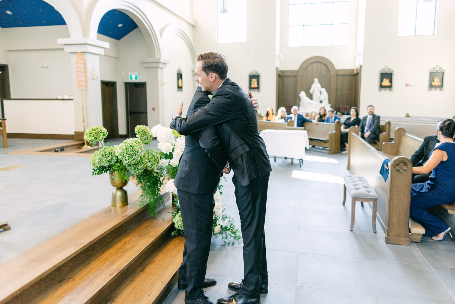 Two people hugging in the aisle of a church during a wedding, with guests seated in pews in the background.