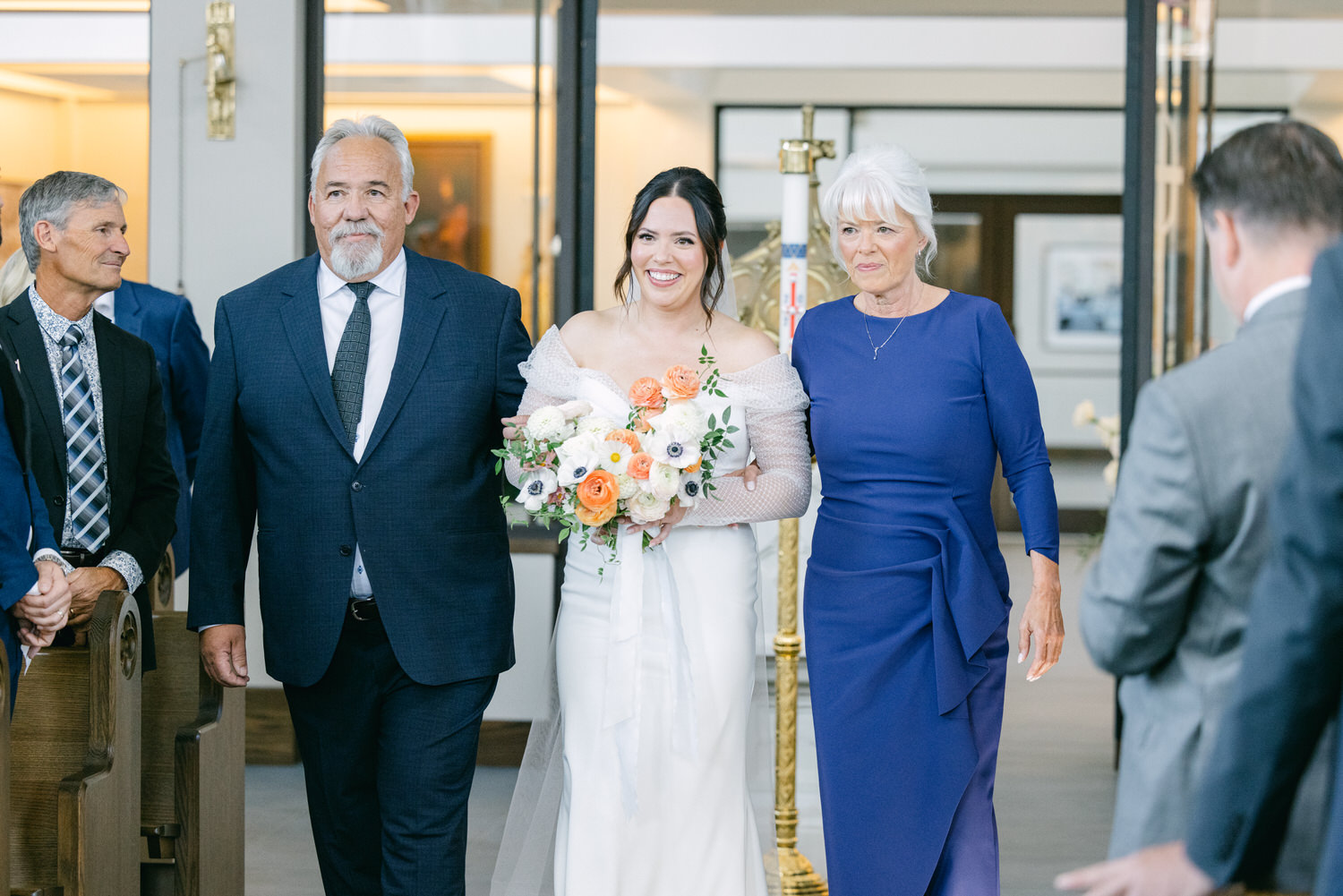 Bride smiling accompanied by parents walking down the aisle at a wedding venue.