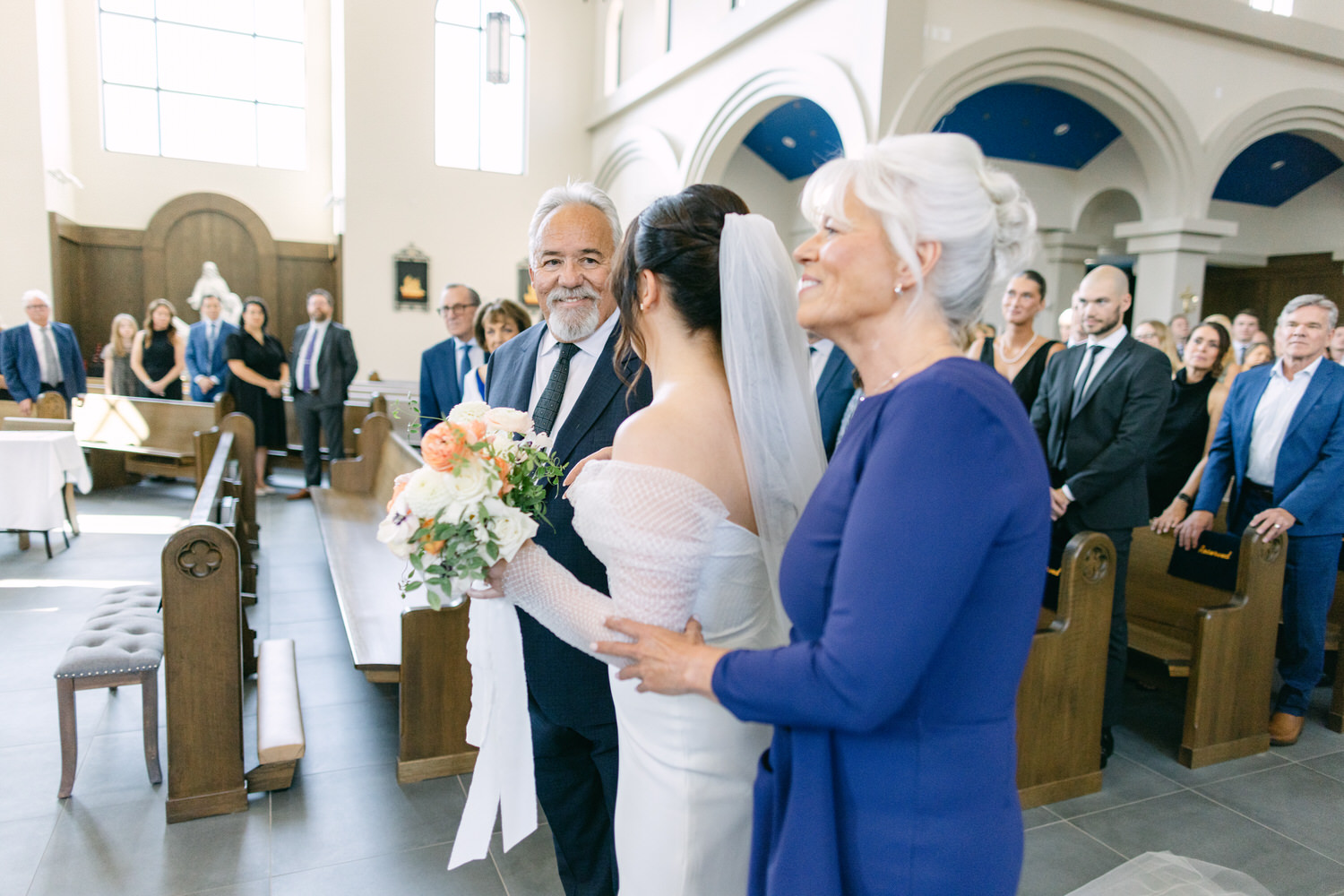 A bride walking down the aisle with an older man and woman, likely her parents, smiling as wedding guests look on in a church setting.