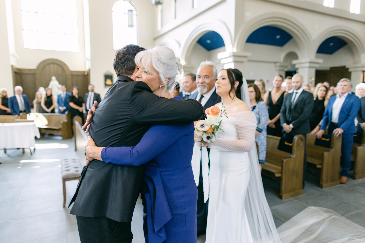 A bride looks on as two guests share a heartfelt hug at a wedding ceremony inside a church.