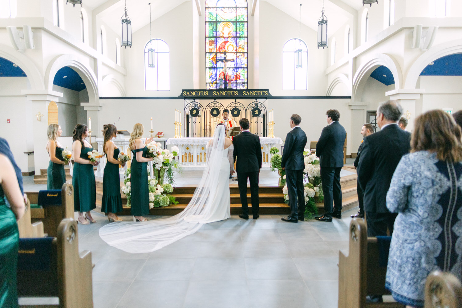 A bride and groom stand at the altar in a church with a priest officiating the wedding, bridesmaids in green dresses to the side, guests in attendance, and a stained glass window in the background.