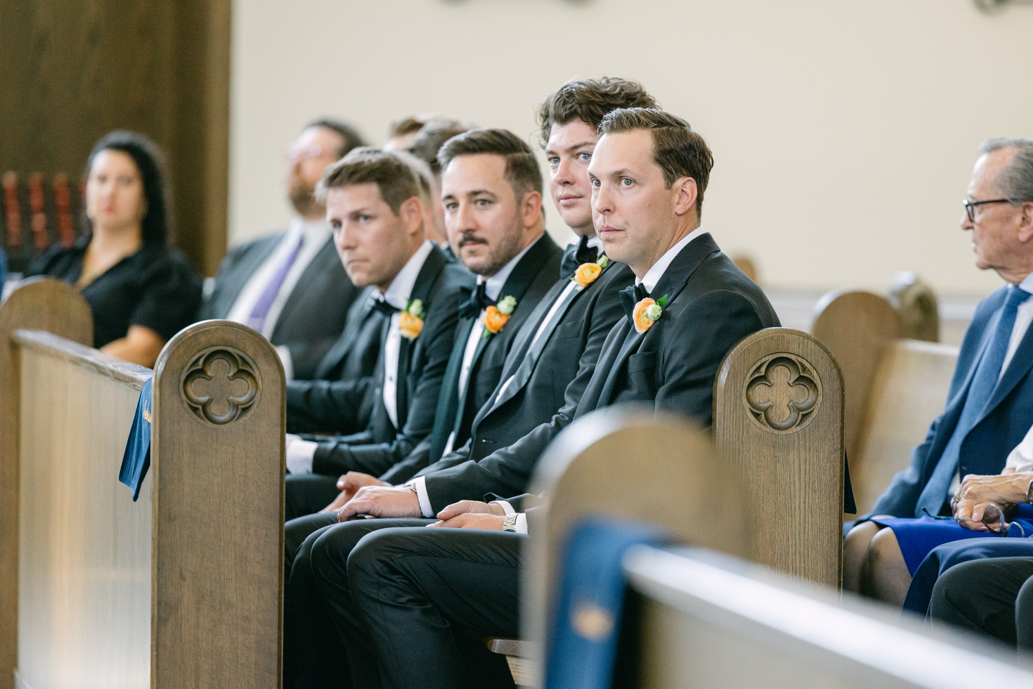 A row of groomsmen wearing black suits with orange boutonnieres sitting attentively in a church during a wedding ceremony.