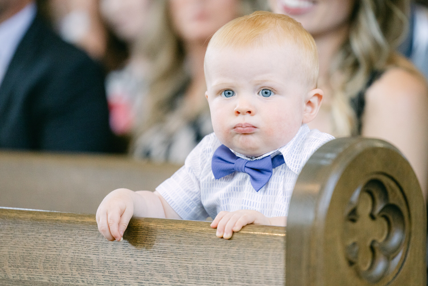 A young child with red hair and blue eyes, wearing a striped shirt and bow tie, peeking over a wooden pew with a curious expression.