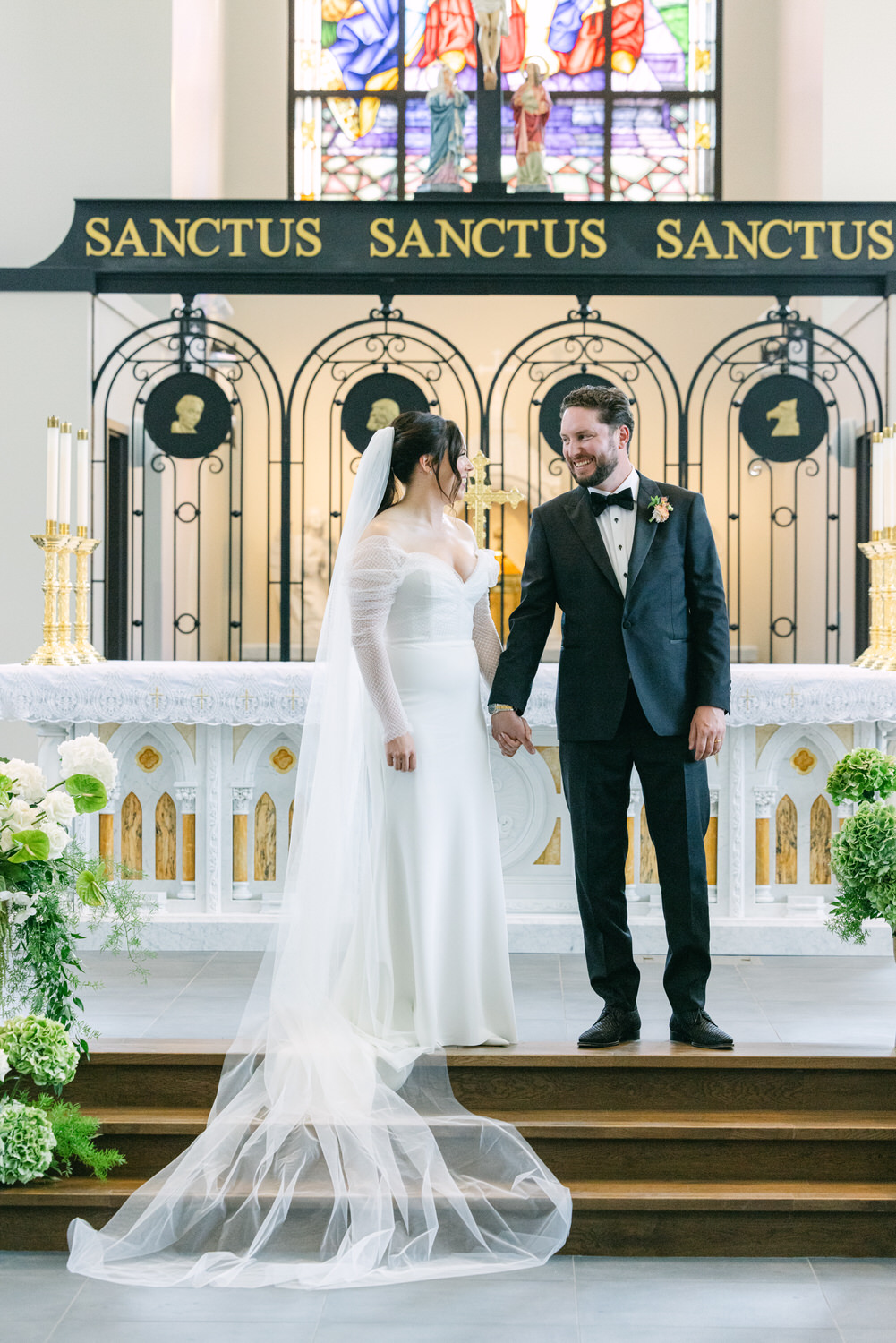 A bride and groom holding hands at the altar of a church, with a background featuring stained glass windows and the word 'SANCTUS'.