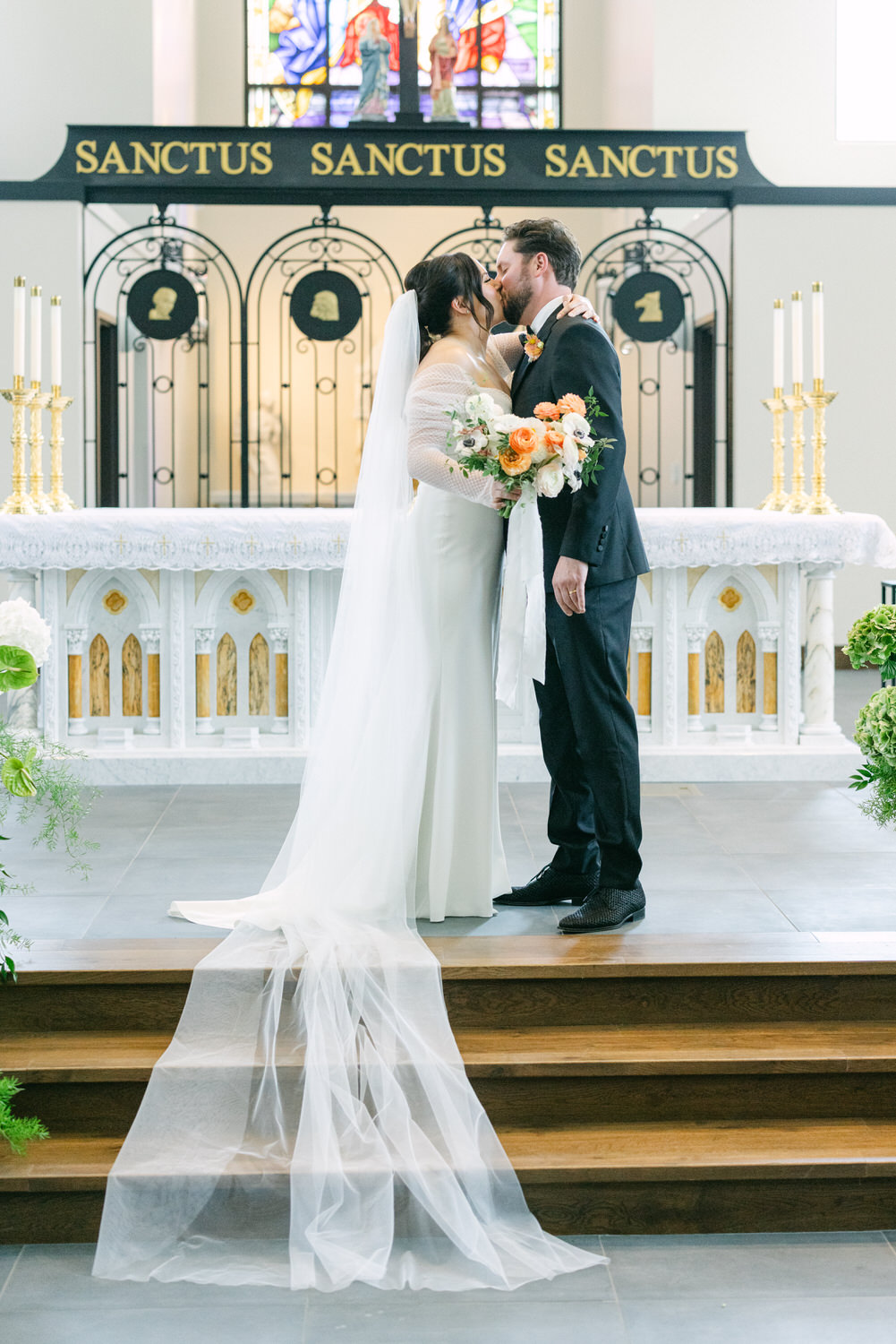 A bride and groom sharing a kiss at the altar with stained glass window in the background, surrounded by candles and wedding decor.