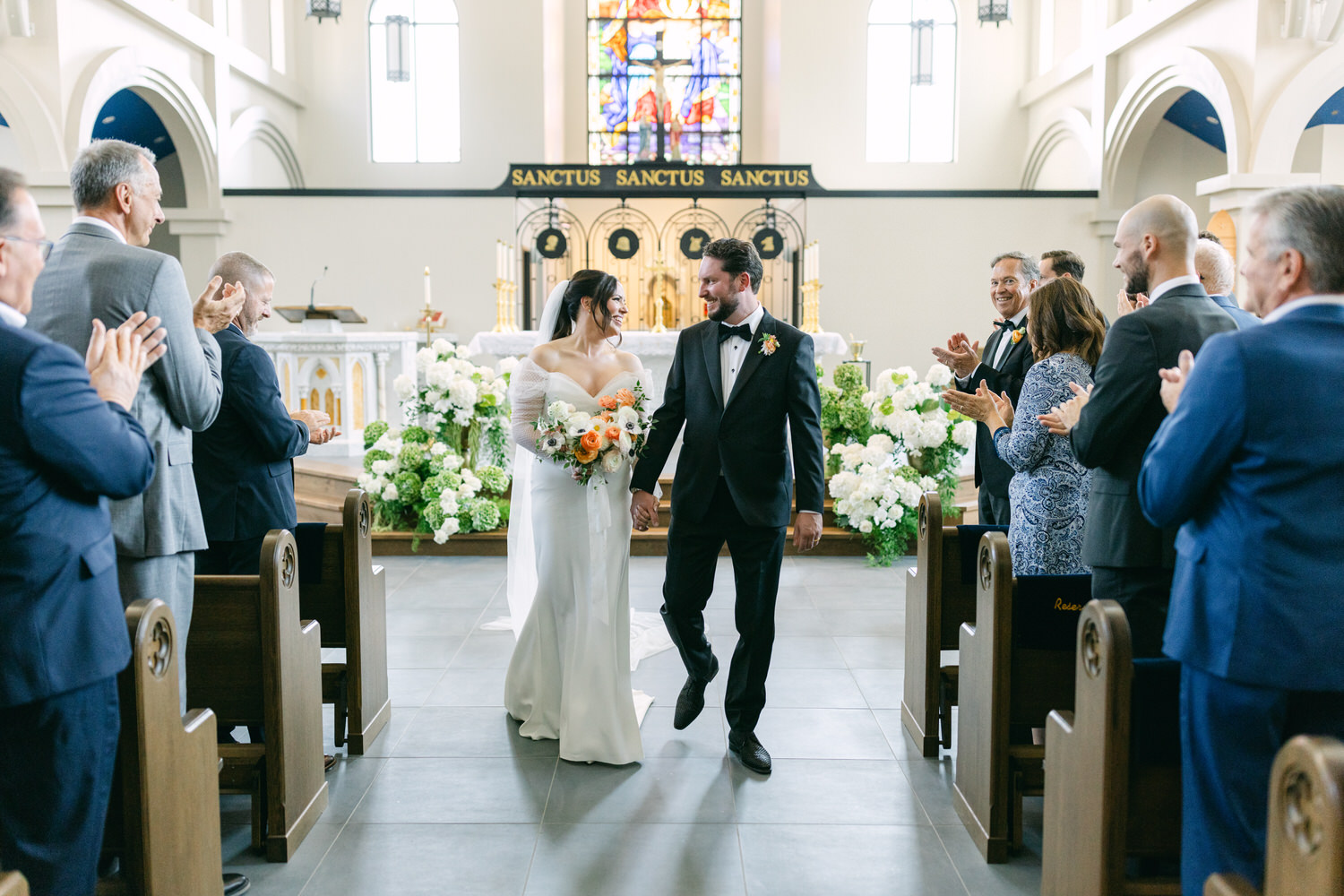 A bride and groom walking down the aisle while wedding guests applaud, in a church with white floral decorations.