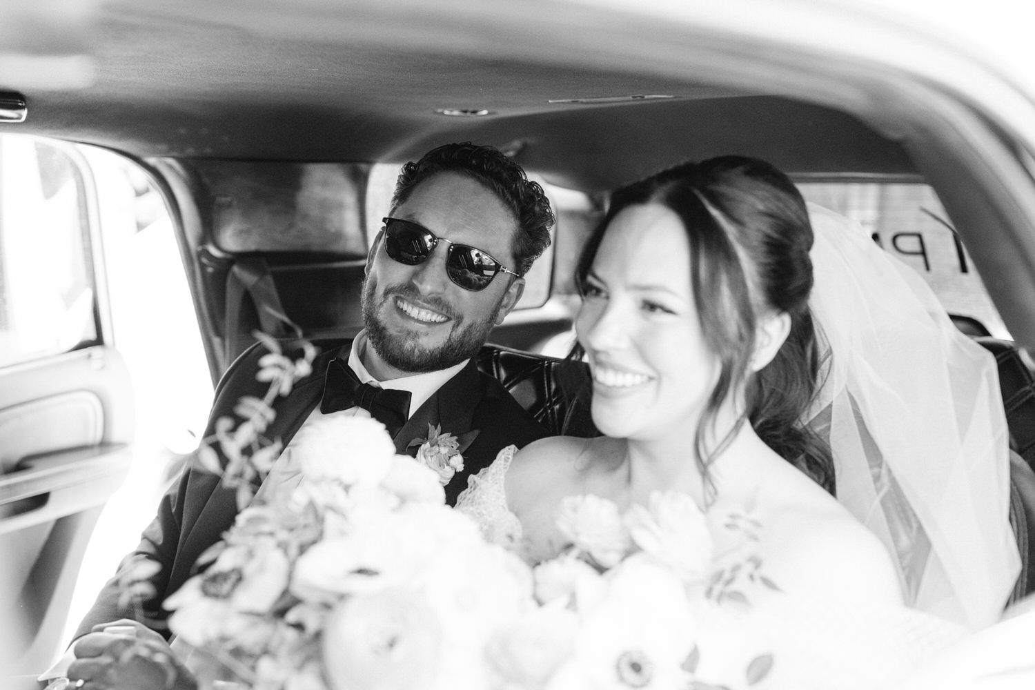 A black and white image of a smiling groom in sunglasses and a radiant bride in a wedding dress, both seated in the back of a limousine with a bouquet of flowers.