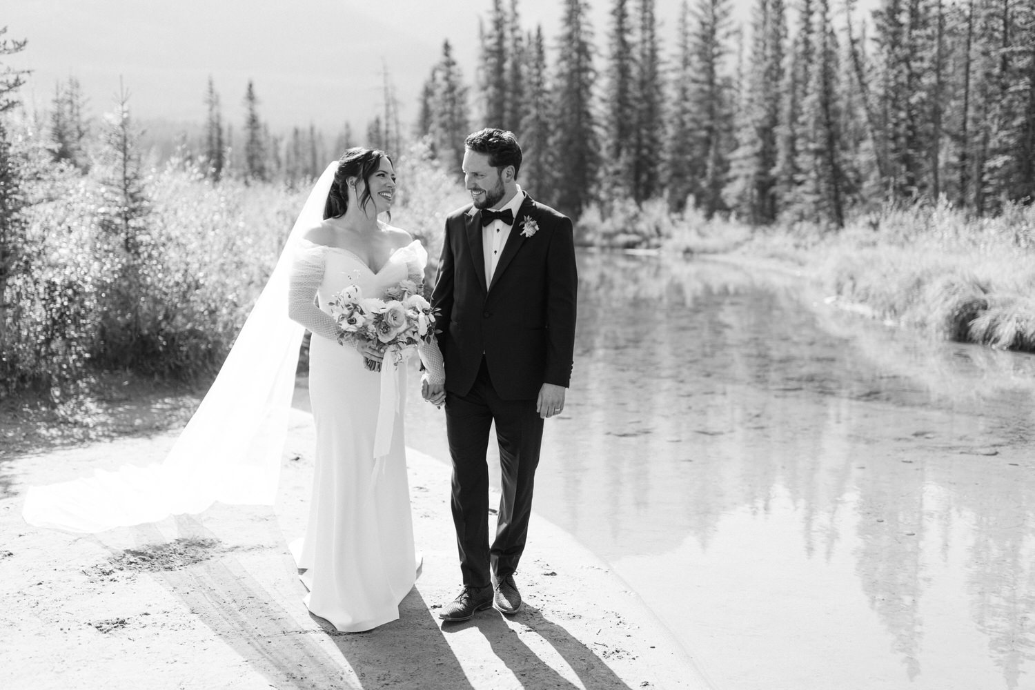 A black and white photo of a bride and groom holding hands and smiling at each other by a tranquil river with forest in the background.