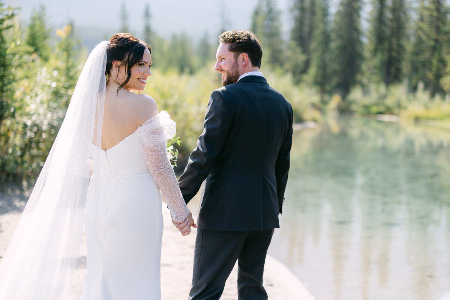 Bride and groom holding hands by a river, with the bride looking back and smiling.