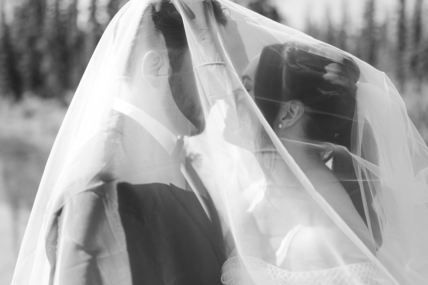 Black and white photograph of a bride and groom close together under a veil with a soft focus forest background.