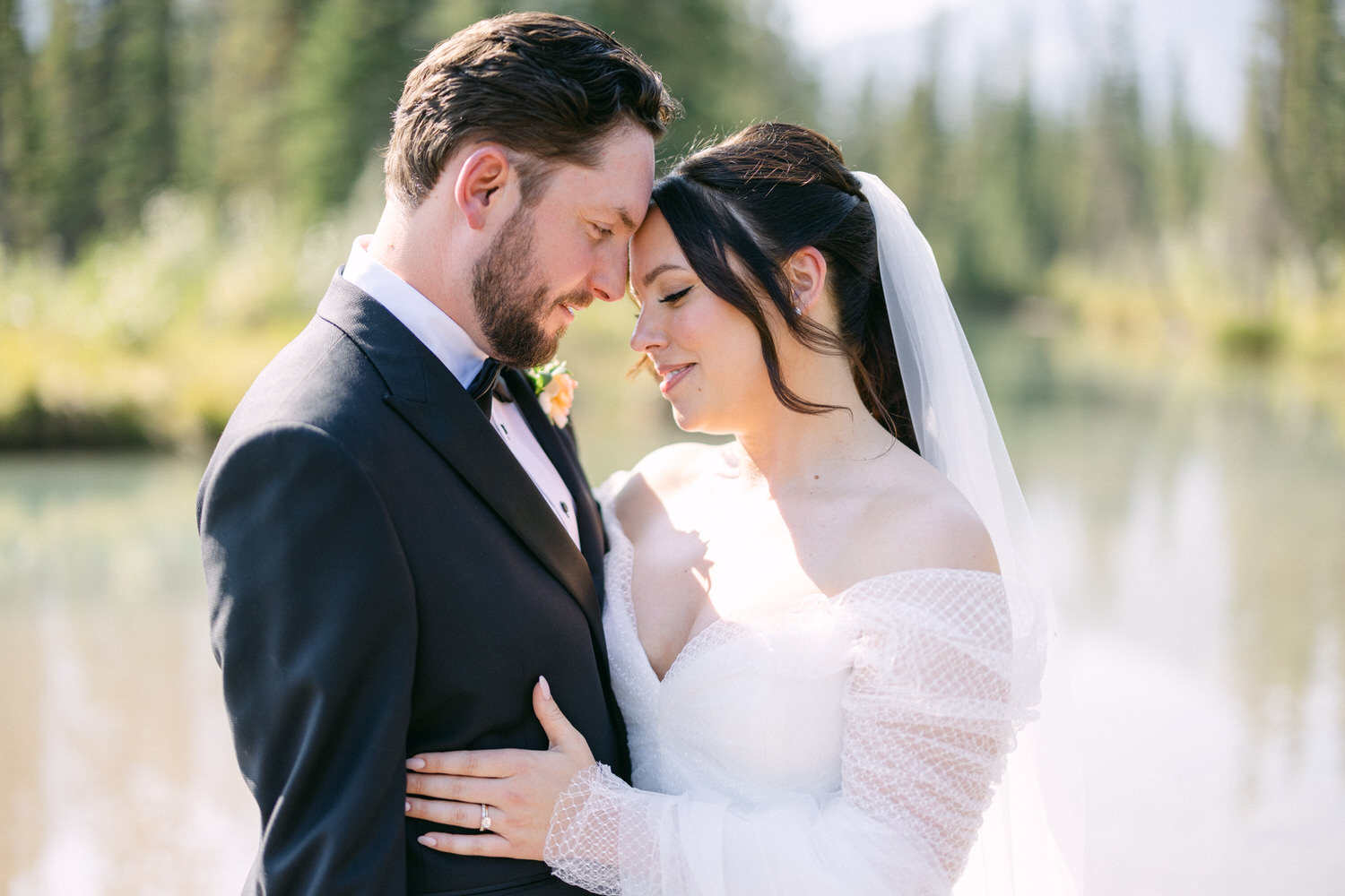 A bride and groom intimately forehead to forehead in their wedding attire with a serene lake and trees in the background.