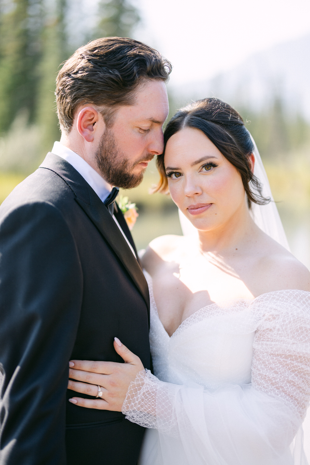 A bride in a white dress and a groom in a black tuxedo intimately embracing, with a soft-focus natural background.