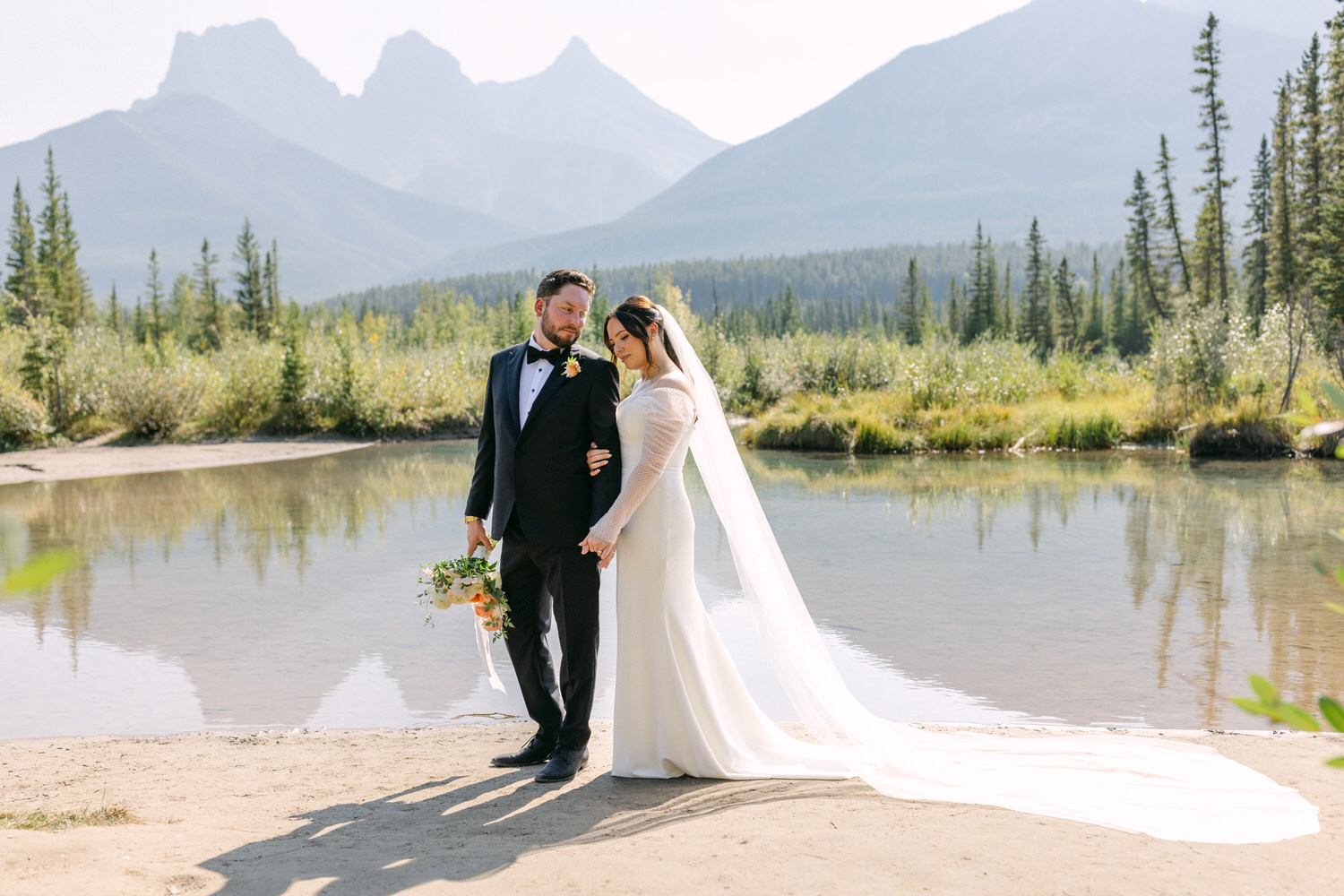 A newlywed couple standing by a lake with mountains in the background, the bride in a long white dress with a trailing veil and the groom in a classic black suit.