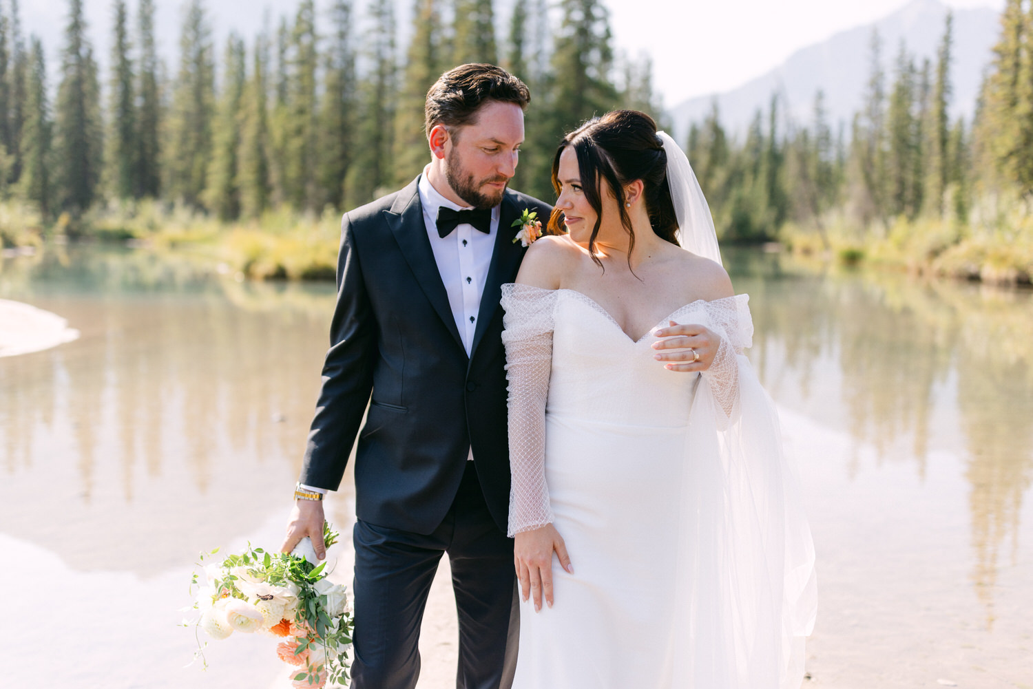 A bride and groom holding hands by a serene mountain lake with pine trees in the background.