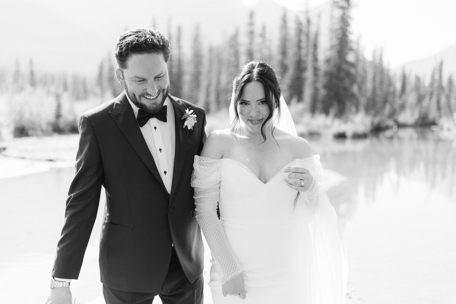 Black and white photo of a smiling bride and groom holding hands, with a serene natural background.