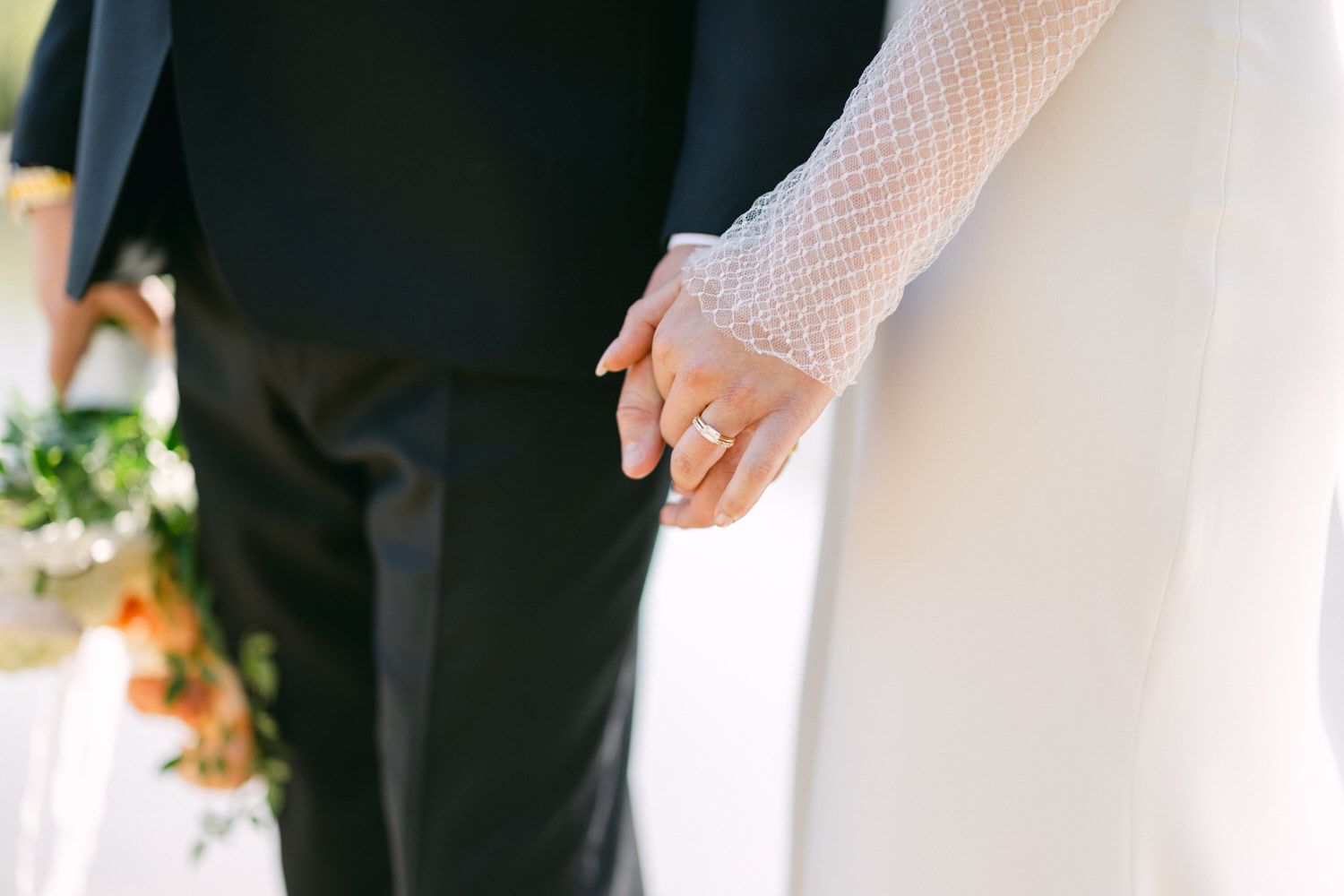 Close-up of a newlywed couple holding hands, with a focus on the wedding rings and the bride's white lace sleeve.