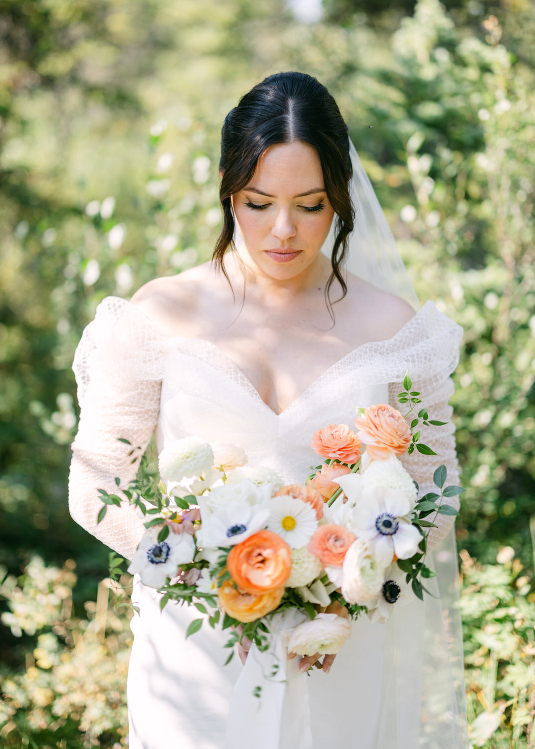 A bride in an off-shoulder wedding dress gazes downward while holding a bouquet of white, orange, and blue flowers, with greenery in the background.