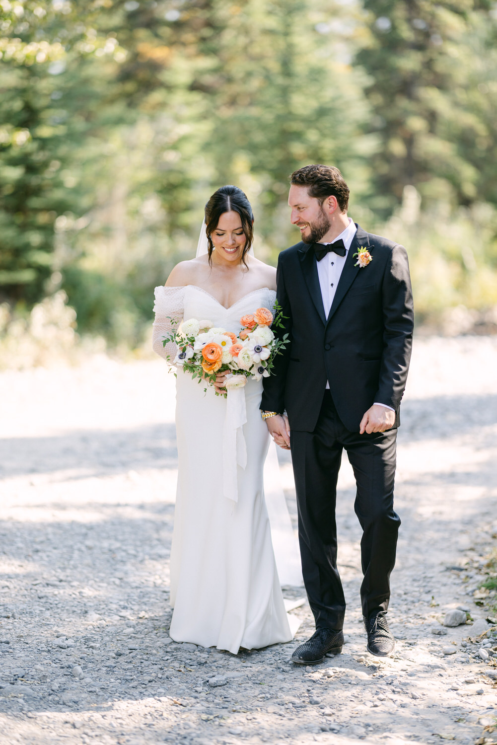 A bride in an off-shoulder wedding dress and a groom in a black tuxedo smiling and holding hands amidst a natural setting.