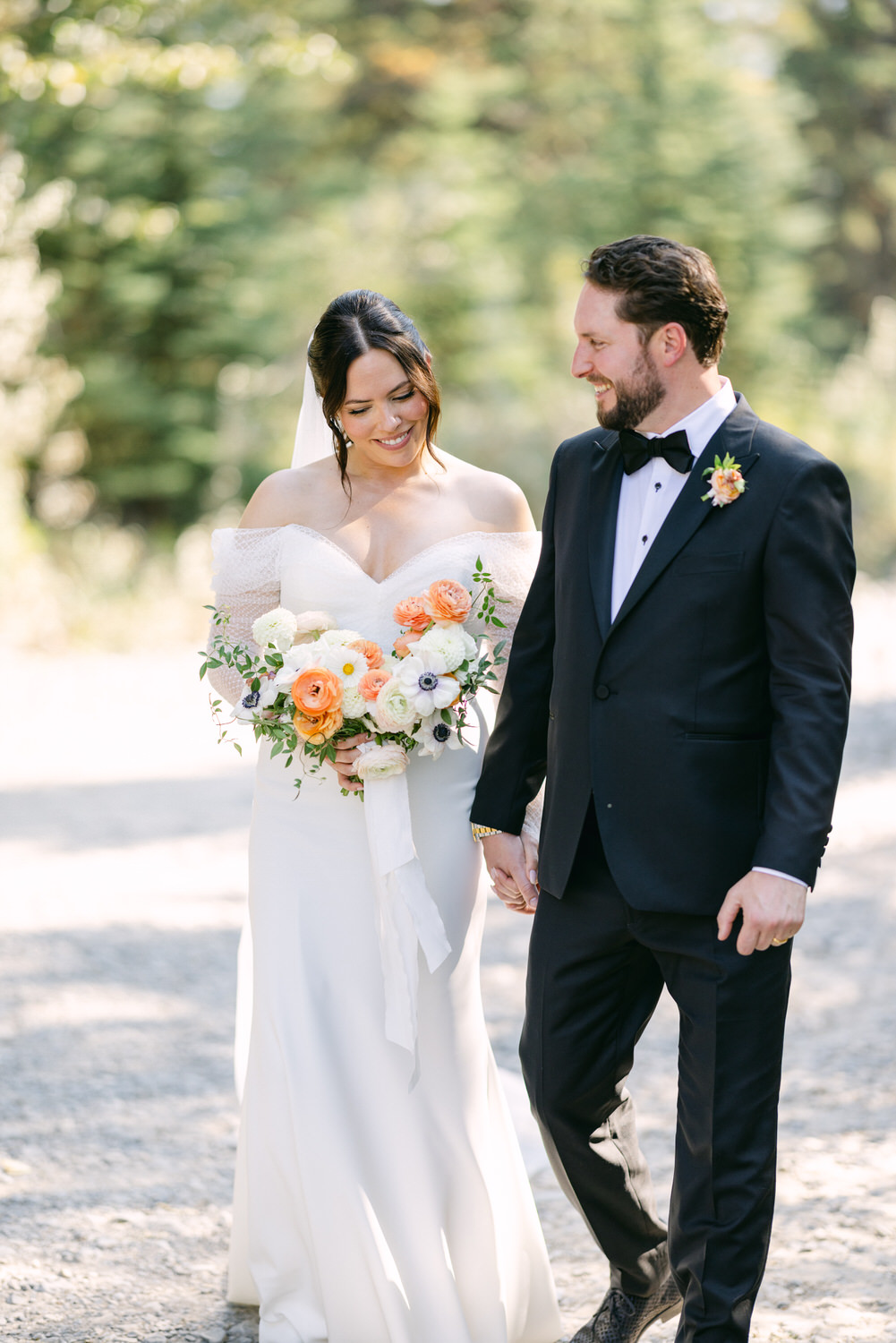 A bride and groom walking hand in hand, the bride holding a bouquet, both smiling and looking down on a sunny day