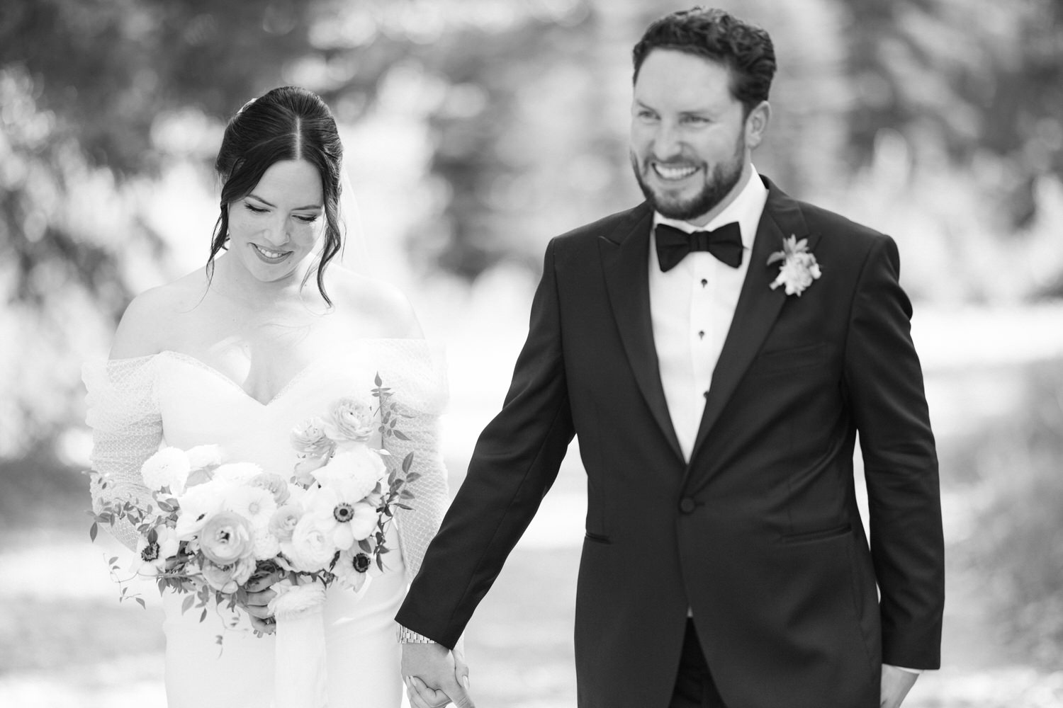 Black and white photo of a smiling bride and groom holding hands while walking, the bride holding a bouquet of flowers.