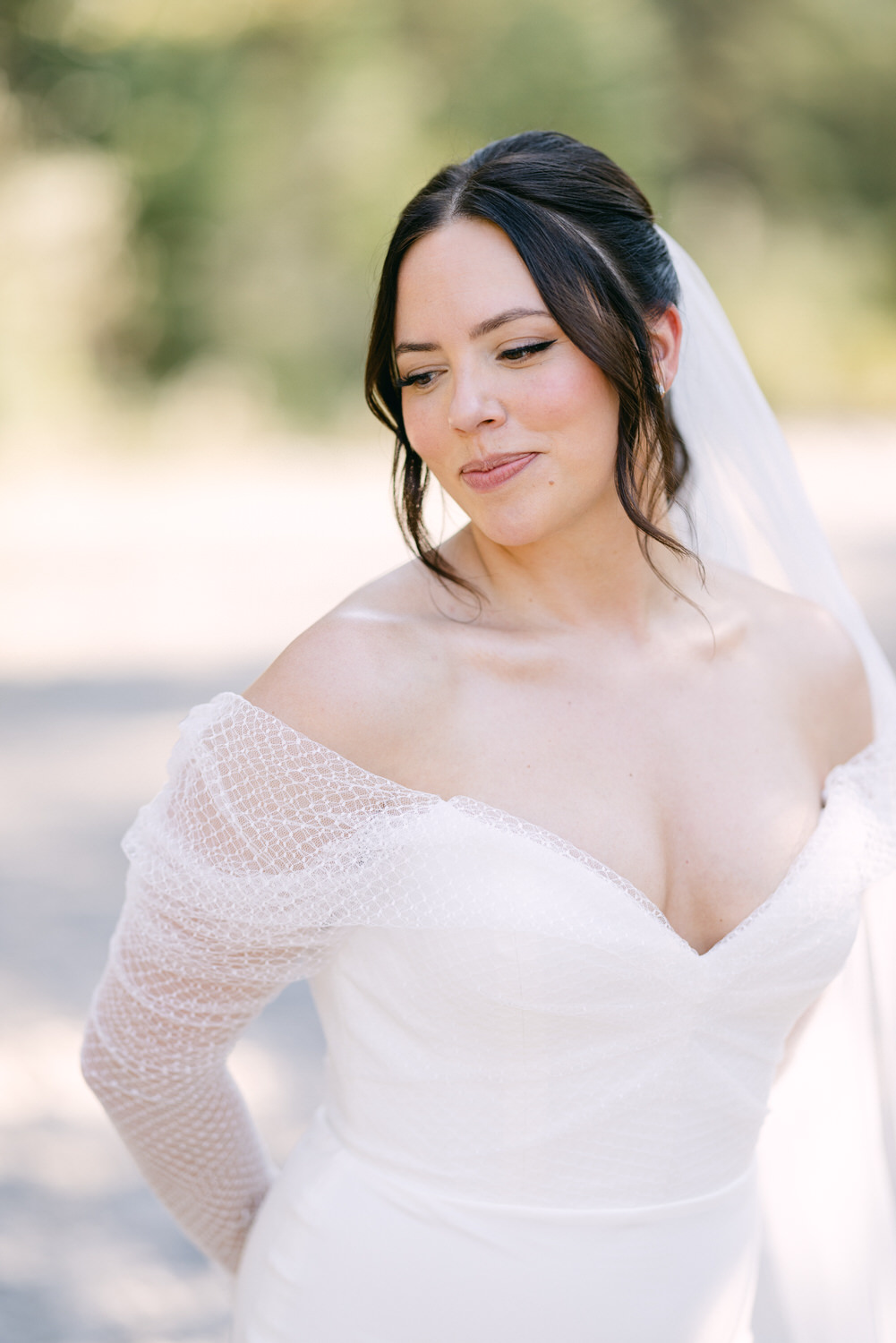 A bride in an elegant white gown with off-the-shoulder lace sleeves and a veil, looking down and slightly smiling.