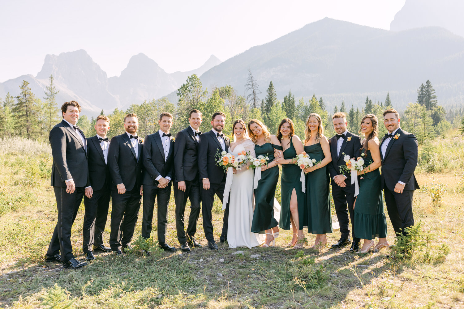 A group of people dressed in wedding attire posing in a natural setting with mountains in the background.