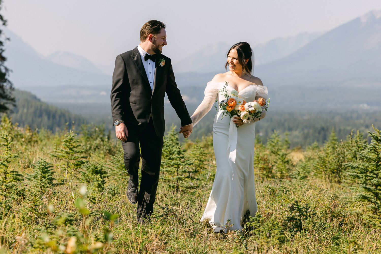 A bride and groom holding hands and smiling at each other in a sunlit mountain meadow, with soft focus mountains in the background.