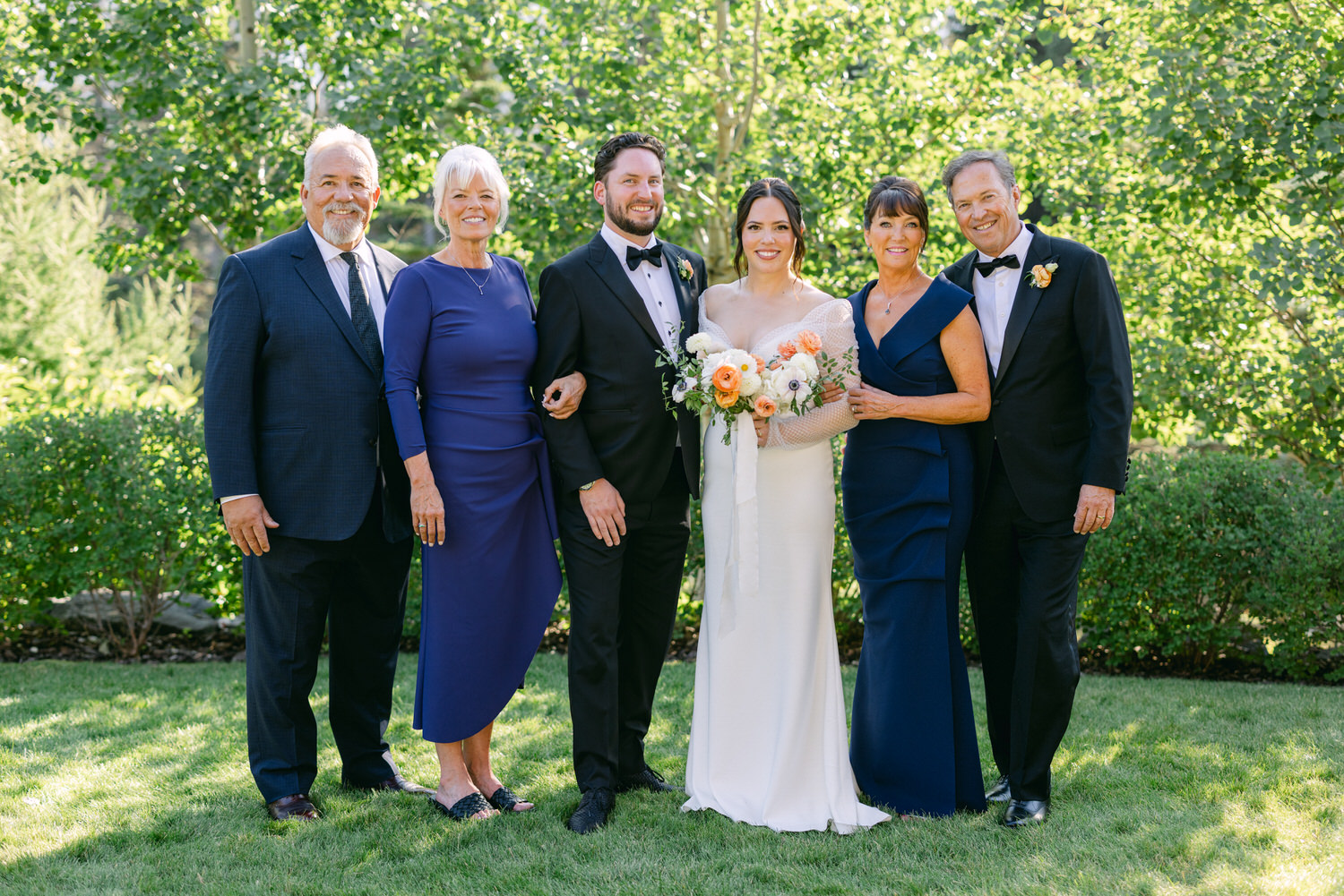 A group of six adults, including a bride and groom, smiling together in formal attire at a garden wedding celebration.