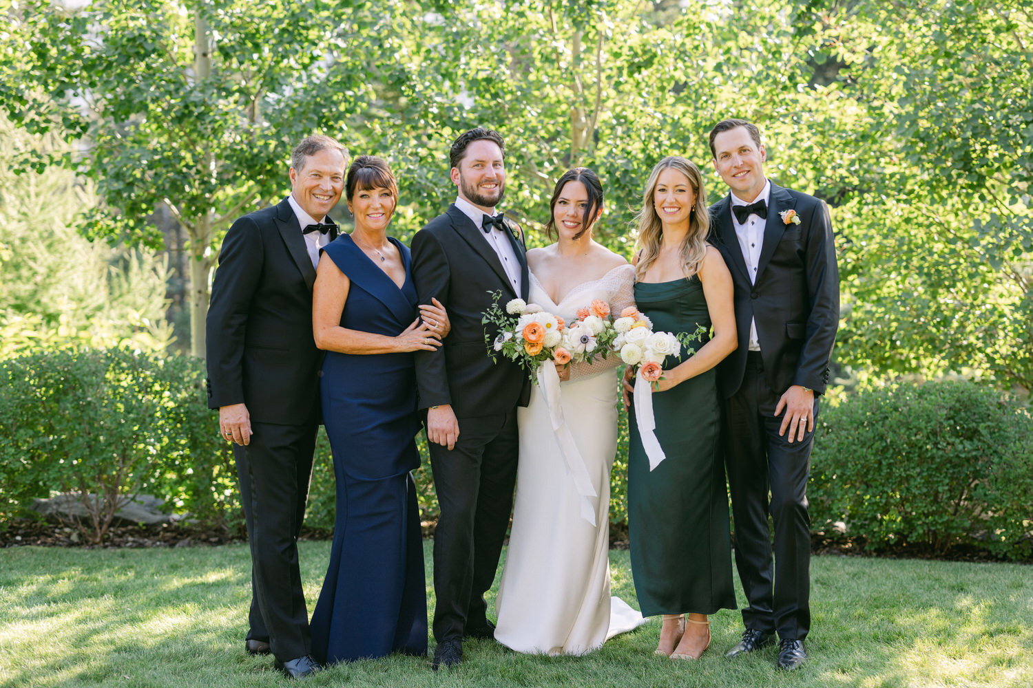 Group of six people dressed in formal attire posing for a photo at a wedding, with a bride and groom in the center, surrounded by lush greenery in an outdoor setting.