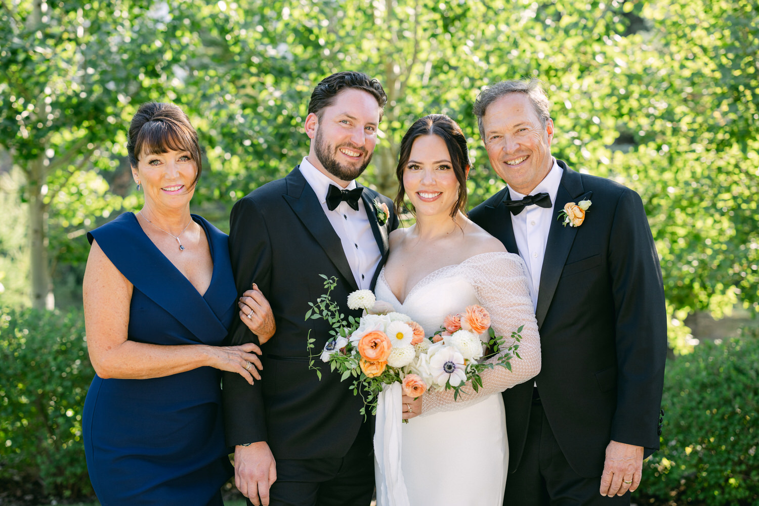 Two women and two men smiling for the camera at a wedding with greenery in the background, one woman holding a bouquet of flowers.