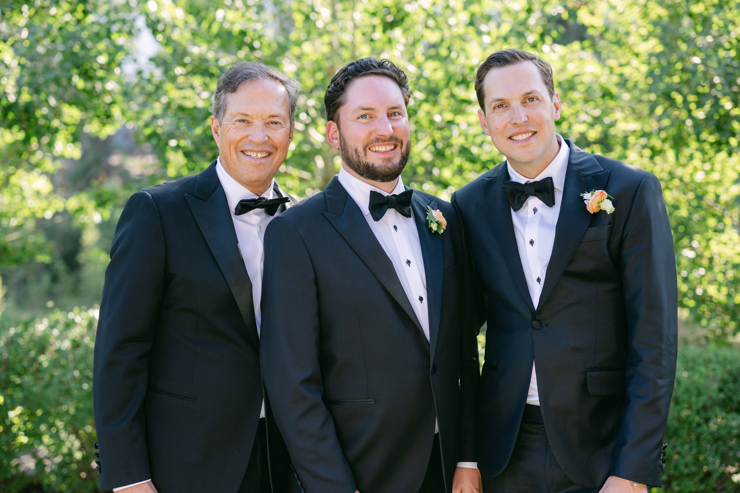 Three smiling men dressed in formal black tuxedos with bow ties and boutonnieres, standing together outdoors with green foliage in the background.