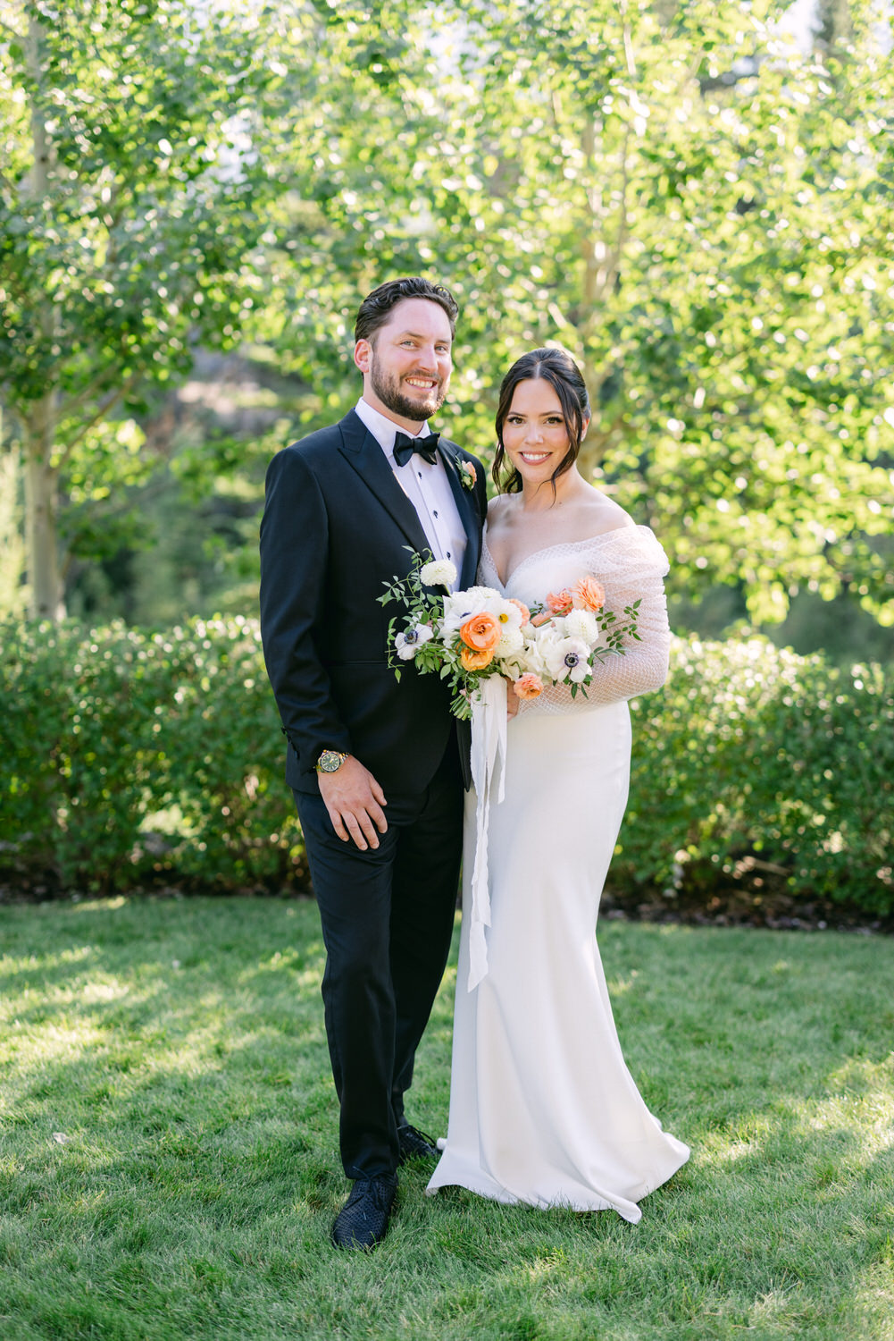 A bride in a white gown and groom in a black suit smiling on a lush green lawn with trees in the background.