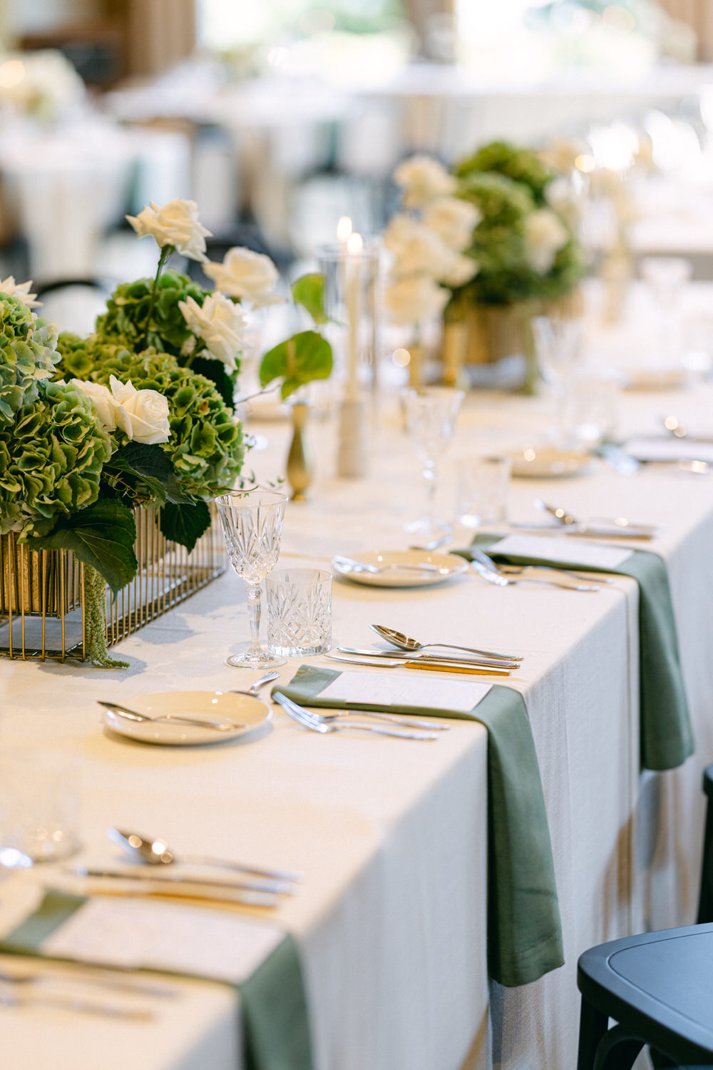 A finely decorated wedding table with white flowers, green hydrangeas, crystal glasses, and gold cutlery on a white tablecloth with a green runner.