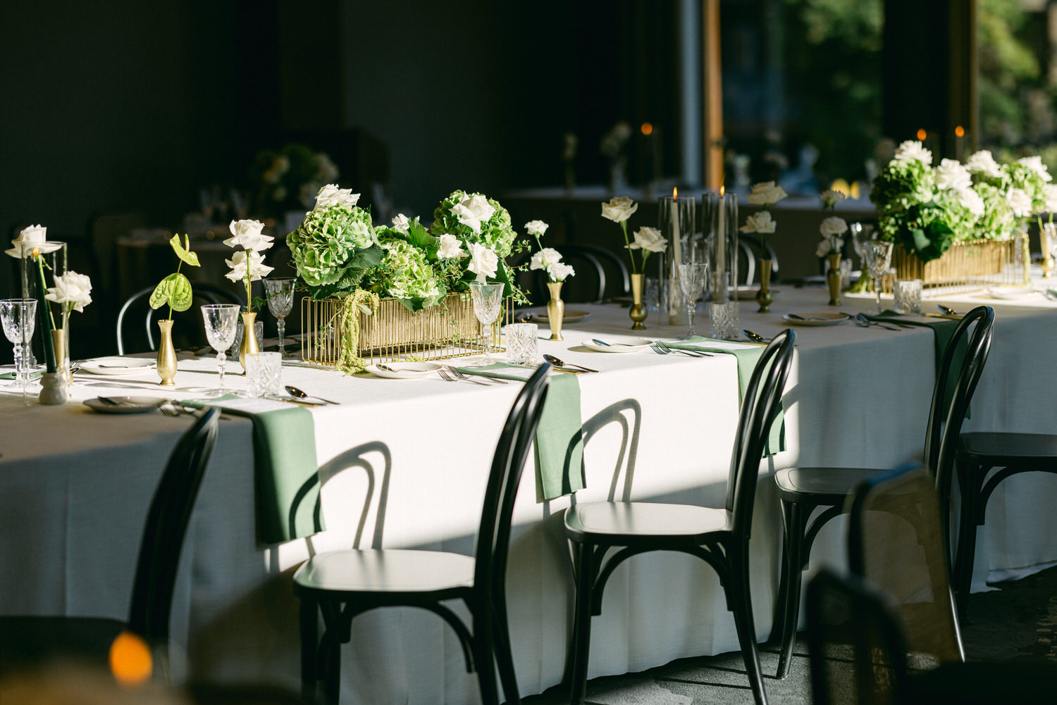 A sunlit elegant dining table setup with green and white floral centerpieces, crystal glassware, and black chairs.