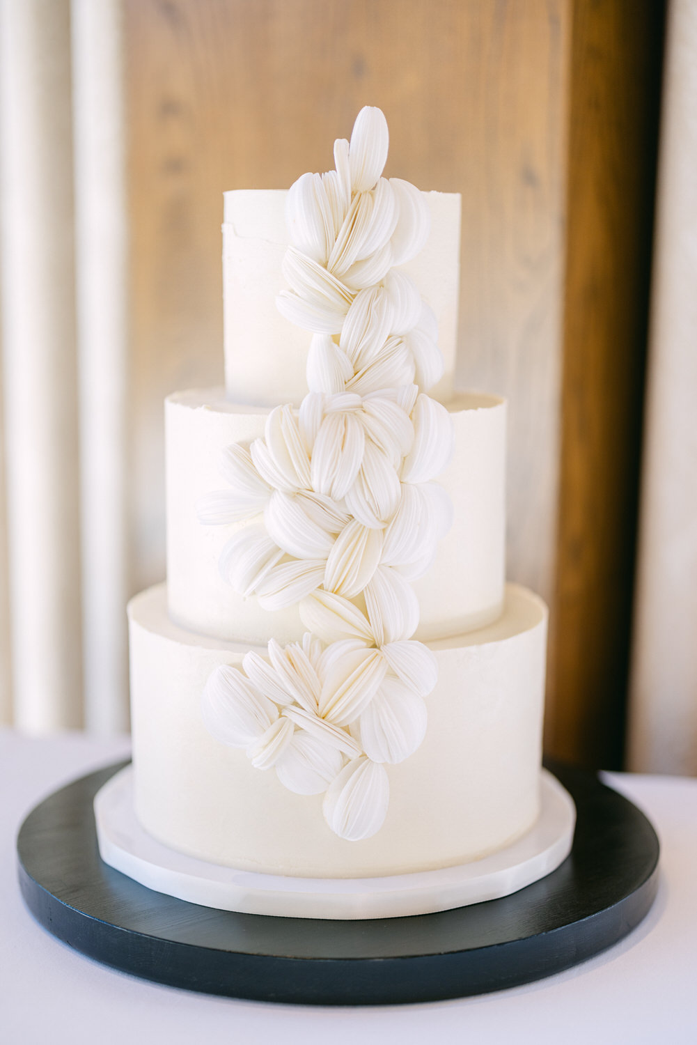 A three-tier white wedding cake decorated with cascading white sugar flowers on a black circular base.