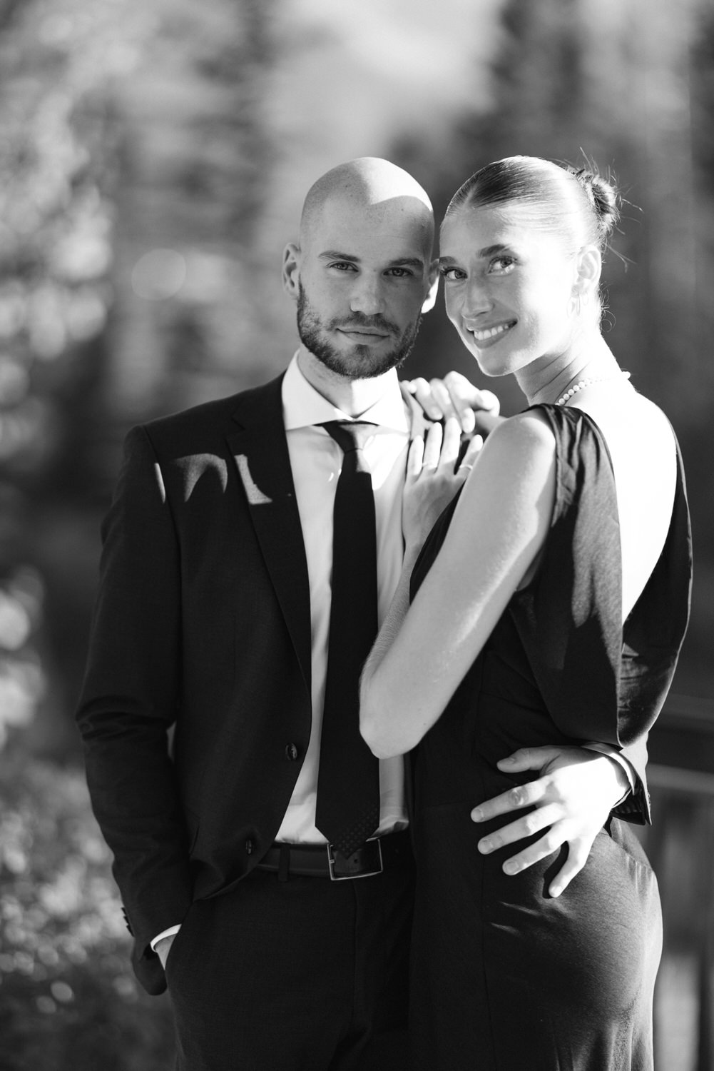 A monochrome photograph of a man and woman dressed in formal wear, the man in a suit and the woman in an evening gown, posing affectionately outdoors.