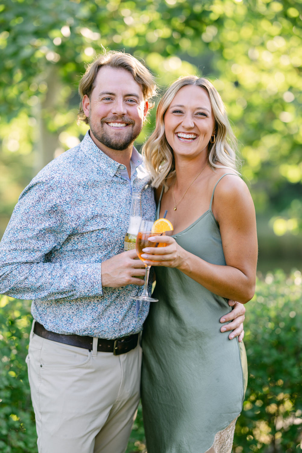 A happy man and woman clinking glasses in a sunny, leafy outdoor setting.