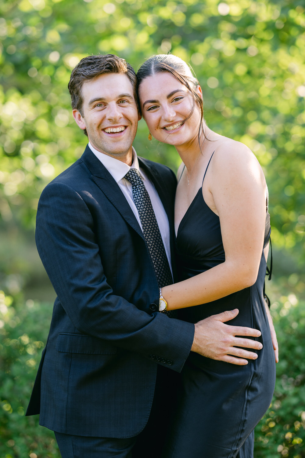 A man in a suit and a woman in a black dress smiling and embracing outdoors with a green, leafy background.