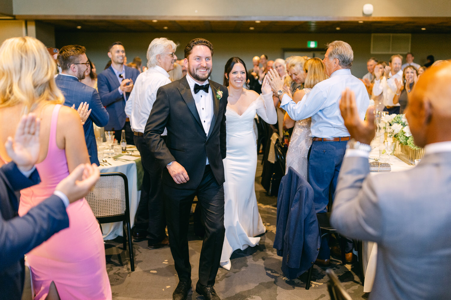 A newlywed couple smiling as they make their entrance to a wedding reception with guests clapping around them.