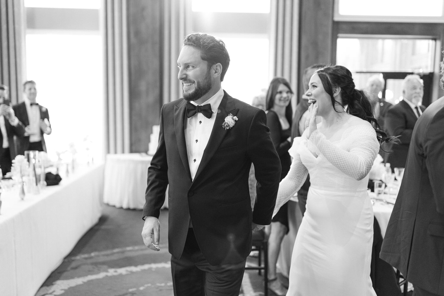 A black and white photo of a smiling bride and groom entering the wedding reception with guests looking on.